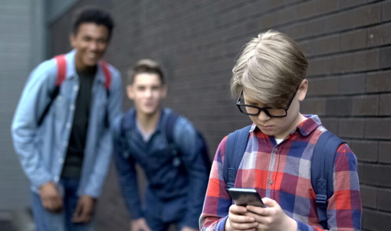 Junior student reading offensive post in phone, boys mock behind, cyberbullying