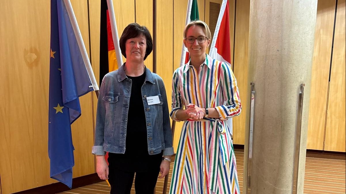 Renate Worms in Jeansjacke und eine Frau im bunten Kleid stehen vor Flaggen der EU, Deutschlands und Italiens