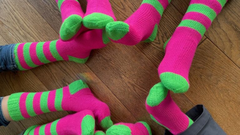 Viele Beine mit pink-grün gestreiften Socken auf einem braunen Holzboden ausgebreitet