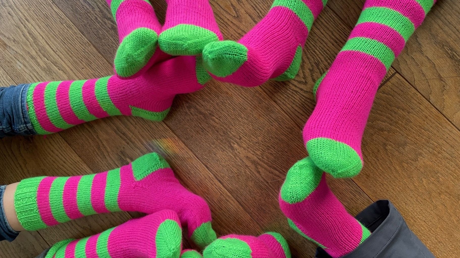 Viele Beine mit pink-grün gestreiften Socken auf einem braunen Holzboden ausgebreitet