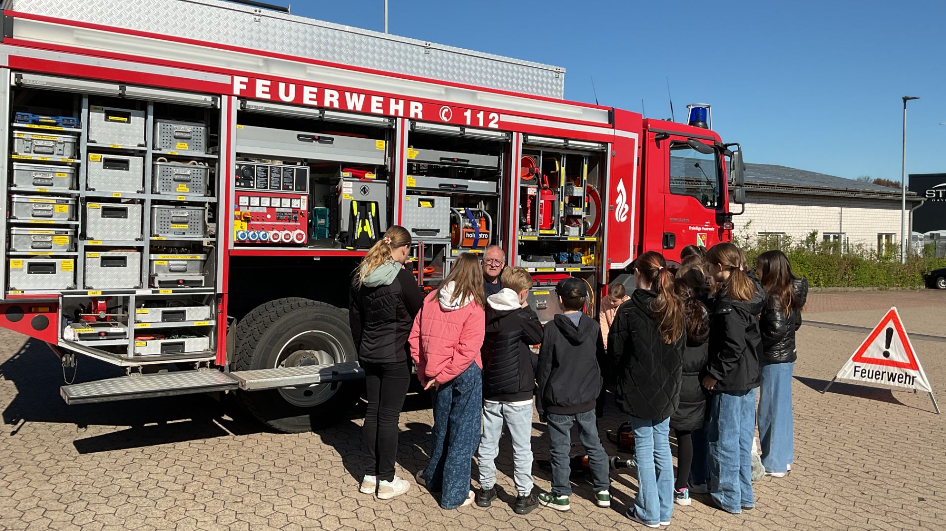 Kindergruppe lernt von Feuerwehrmann an einem roten Feuerwehrauto mit offenem Geräteraum draußen auf einem Hof