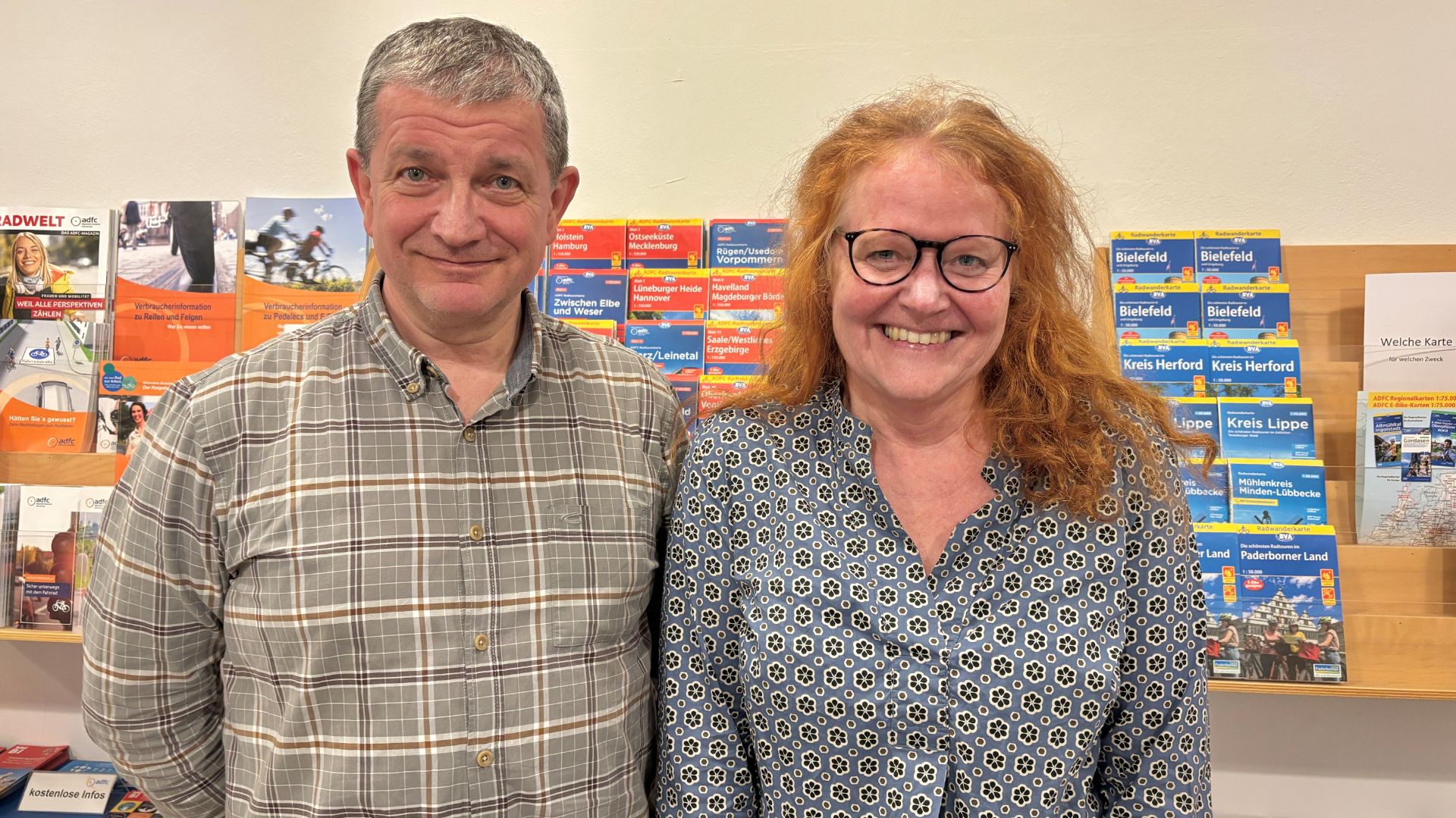 Ruth Hachmann und Bernd Diekmann lächeln vor einem Regal mit ADFC-Radwanderkarten und Broschüren in Bielefeld