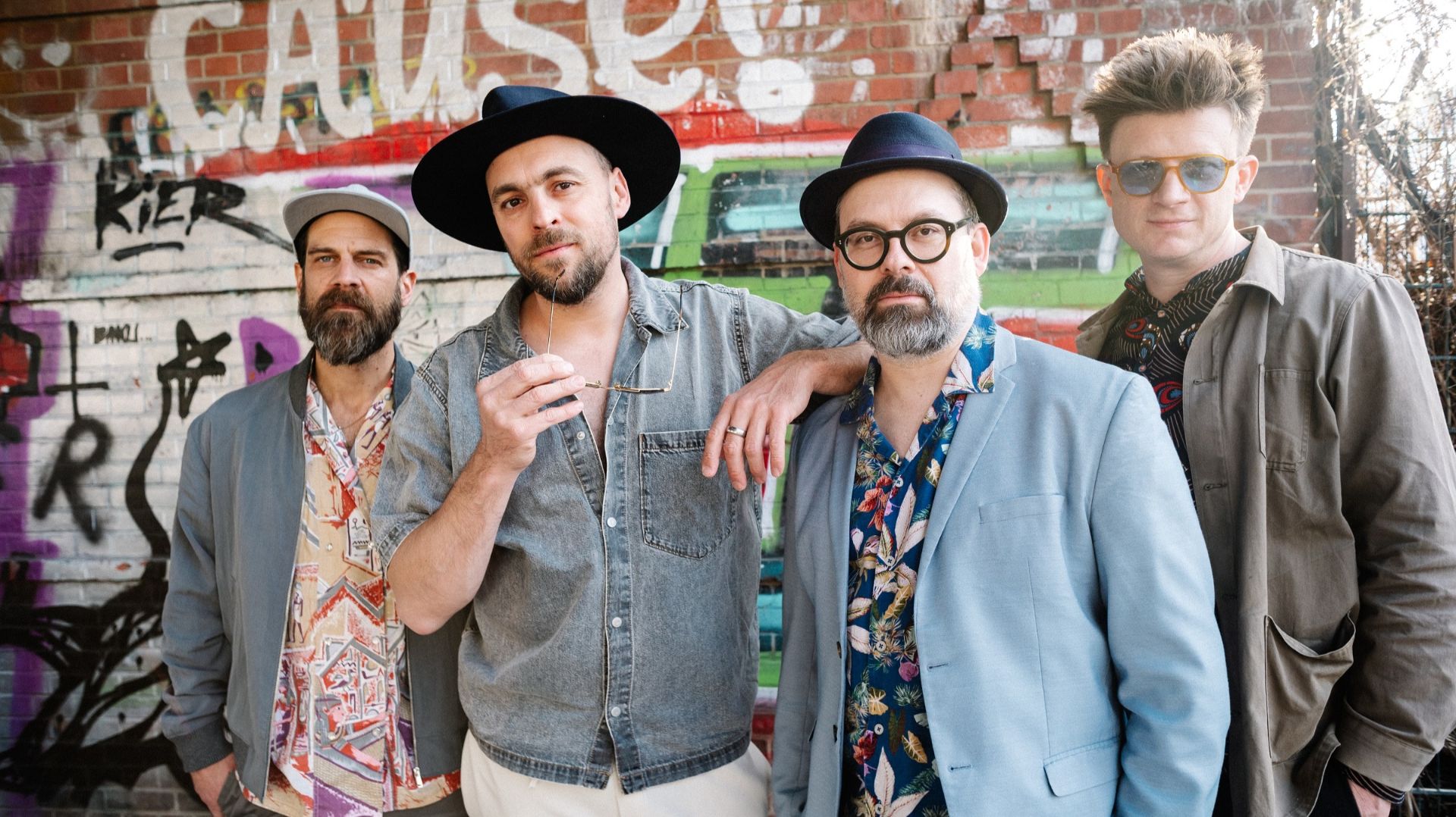 Max Mutzke (Hut) und Matti Klein (Brille) mit zwei weiteren Musikern vor Graffiti-Ziegelwand blicken in Kamera