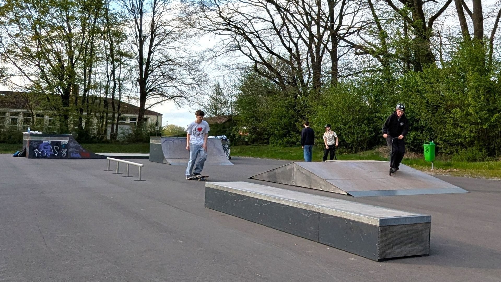 Jugendliche skaten und scootern in einem Skatepark mit Rampen, Boxen und Rails, umgeben von Bäumen und einem Gebäude