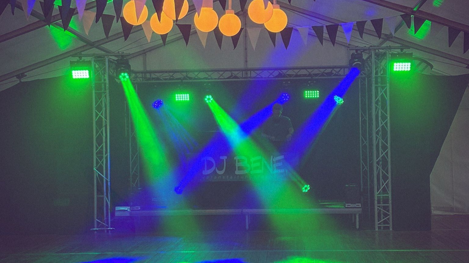 Eine DJ-Bühne mit "DJ BENE"-Schriftzug, grünen und blauen Lichtern in einem Partyzelt mit Girlanden und Kugelleuchten