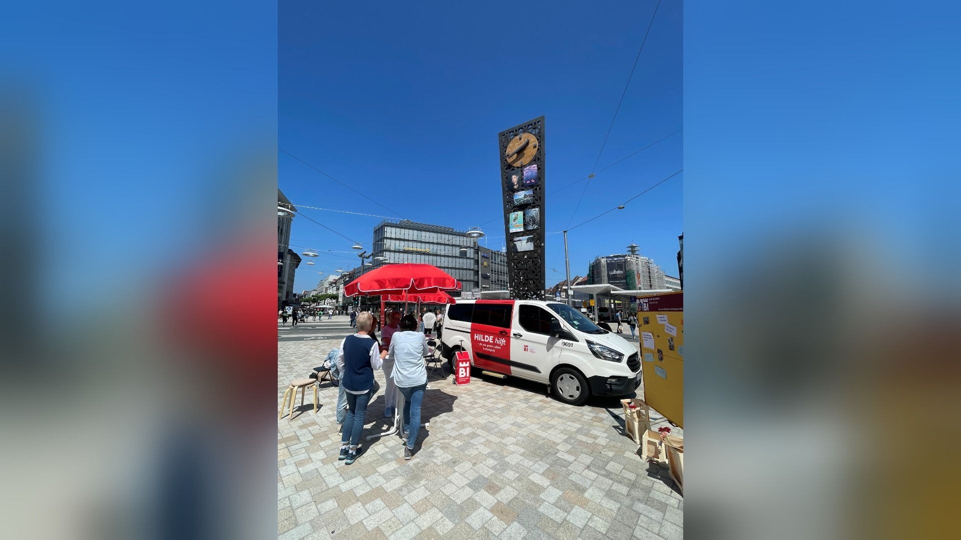 Aktionstag auf einem Platz mit "HILDE hilft" Van, rotem Schirm, Menschen und hoher Uhr-Säule im Hintergrund