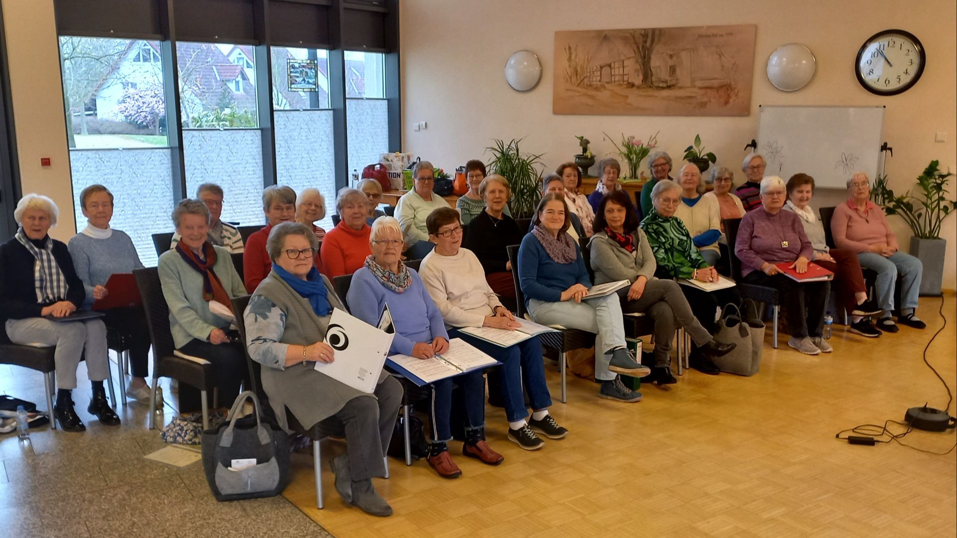 Eine Gruppe von Frauen sitzt in Reihen in einem hellen Raum mit großen Fenstern und Holzboden, aufmerksam nach vorne
