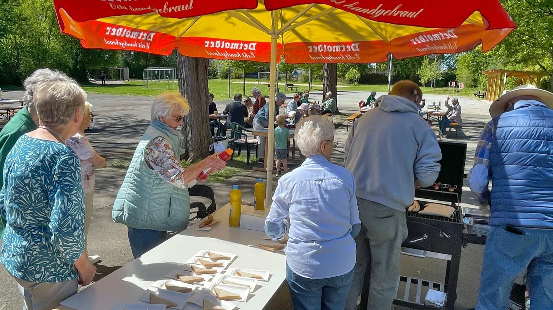 Viele Menschen auf einer Maifeier versammeln sich um einen Grillstand mit Würstchen und Soßen unter einem großen