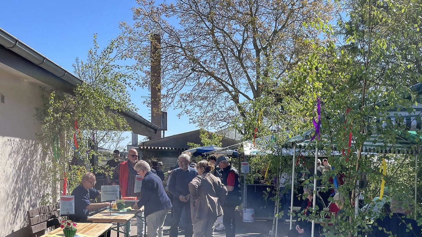 Besucher bei einem sonnigen Maifest unter mit bunten Bändern geschmückten Bäumen und Festzelten, Wertmarkenverkauf