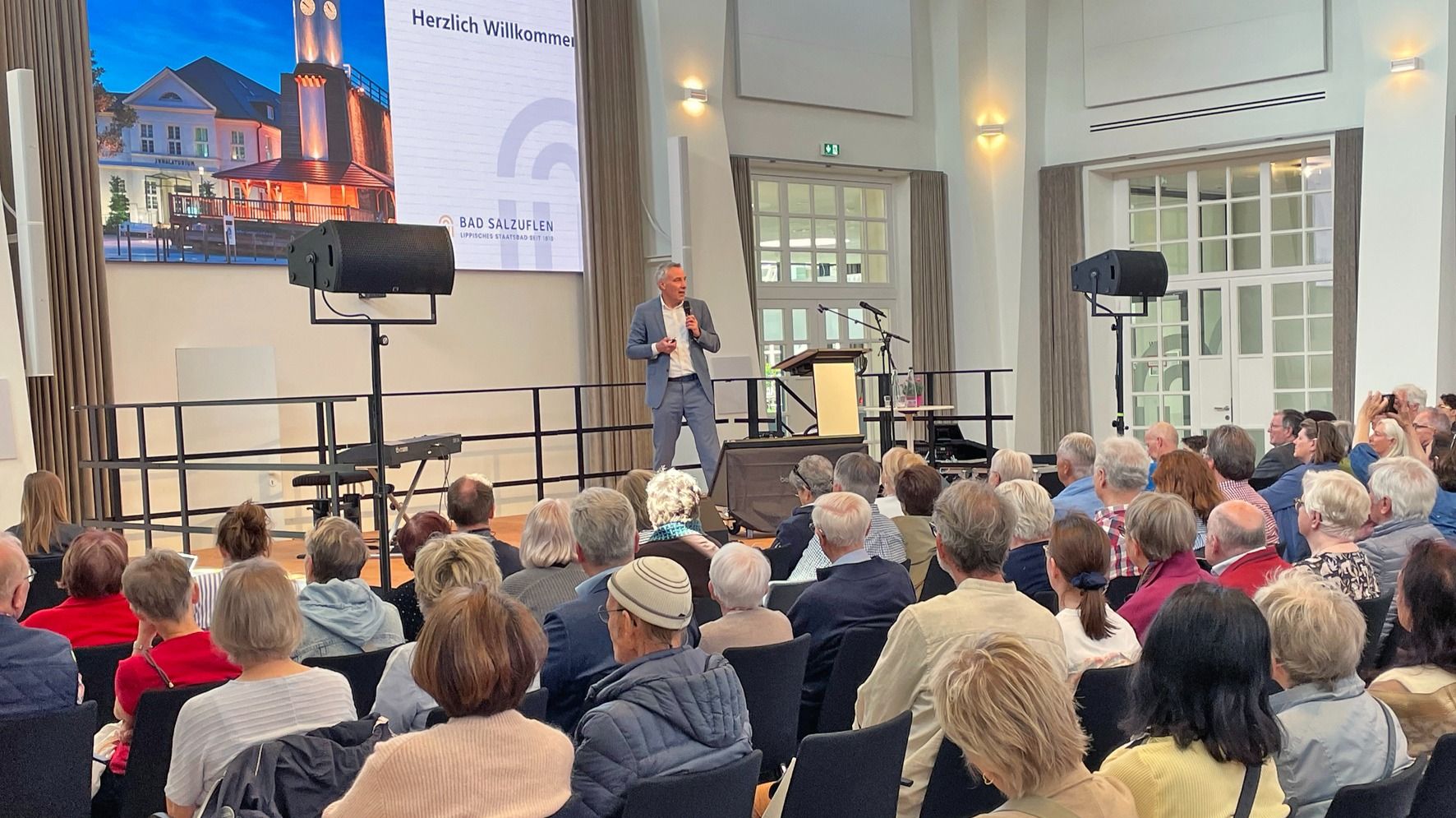 Mann im Anzug hält Rede auf einer Bühne vor einem sitzenden Publikum, hinter ihm Leinwand mit Gebäude und Bad Salzuflen