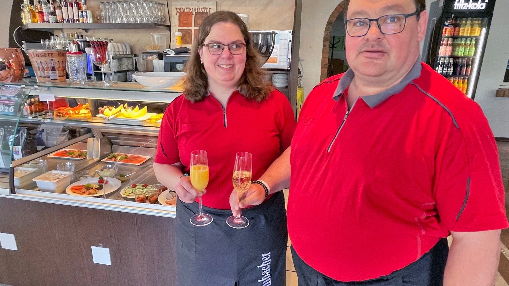 Ein Mann und eine Frau in roten Poloshirts halten Sektgläser vor einer Theke mit Speisen und Getränken