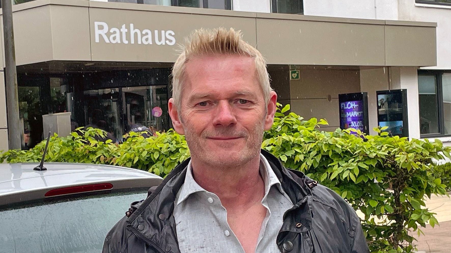Frank Elbrächter mit blonden Haaren und Dreitagebart lächelt vor dem Rathaus mit Büschen und einem Auto