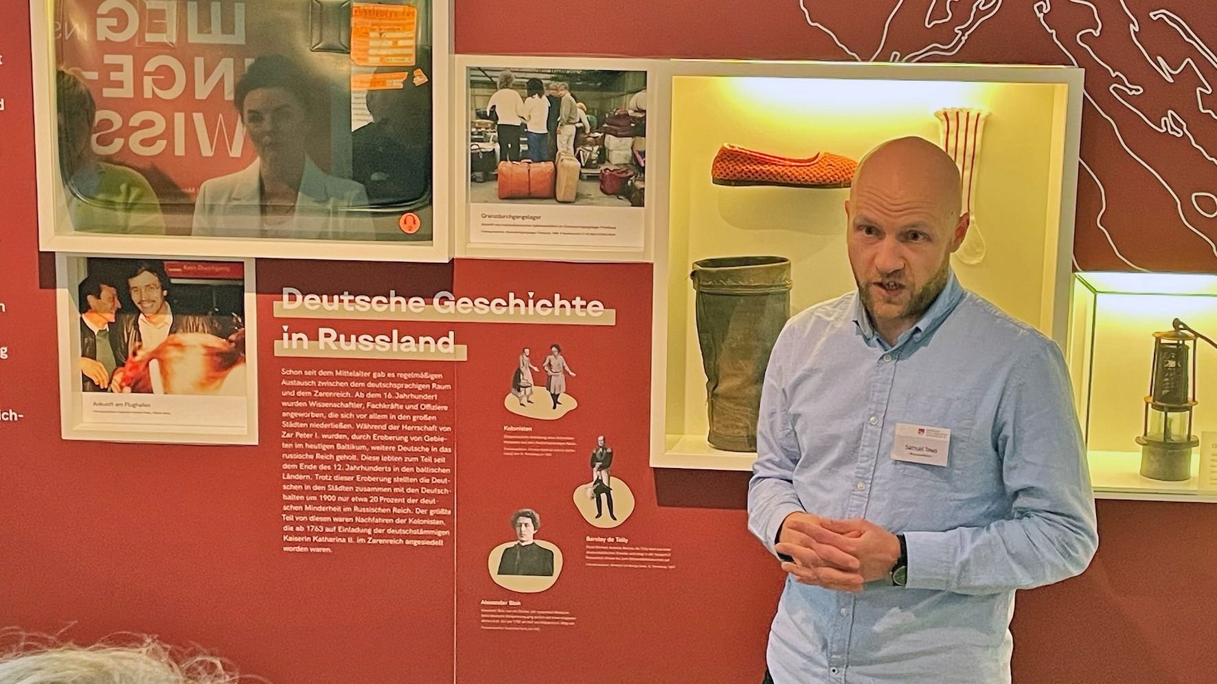 Ein Mann mit Bart und hellblauem Hemd spricht vor einer Museumswand mit Exponaten zur russlanddeutschen Geschichte