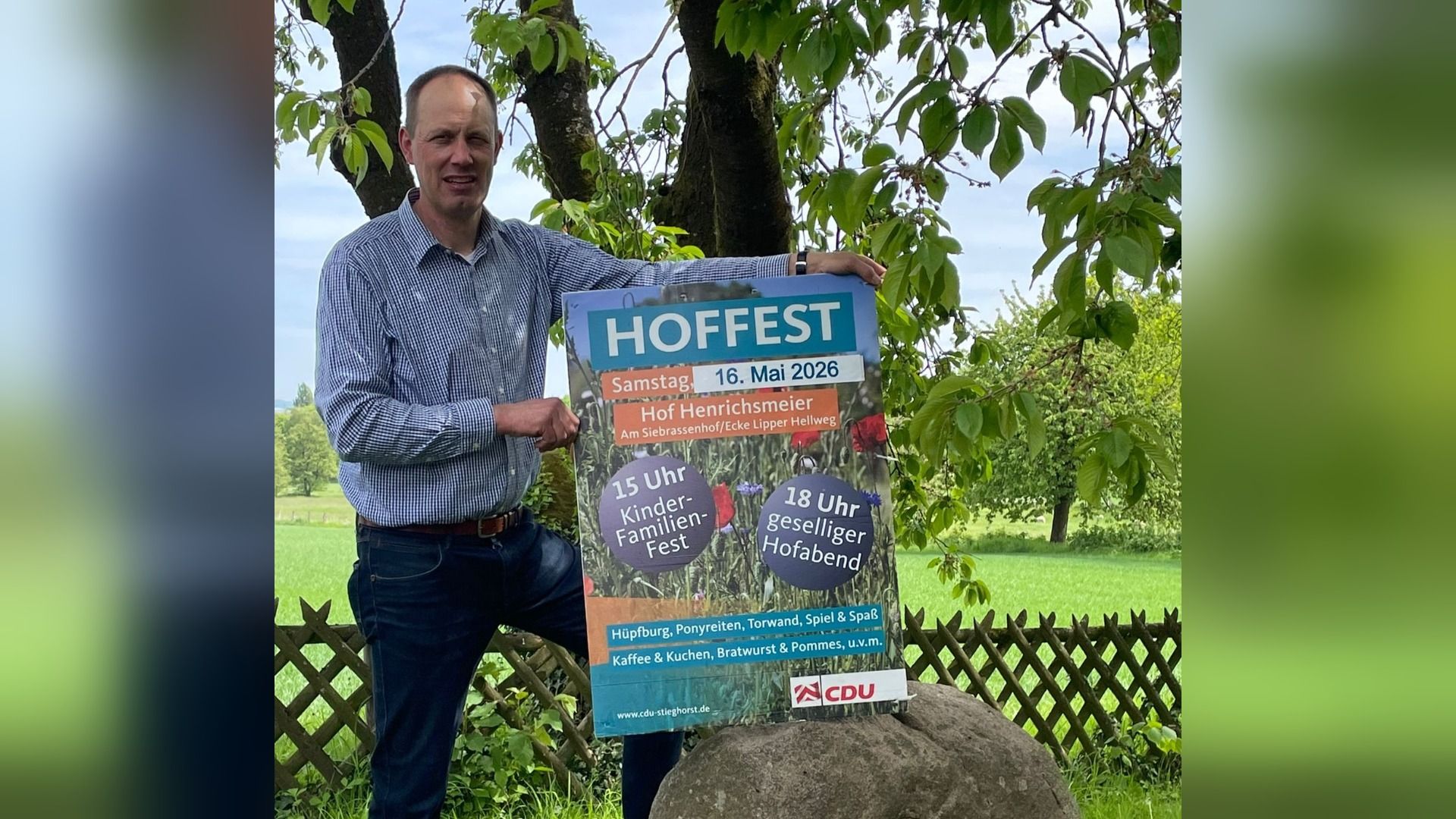Mann im Freien hält ein großes Plakat für ein Hoffest am 16. Mai 2026, das ein Kinderfest und einen Hofabend ankündigt