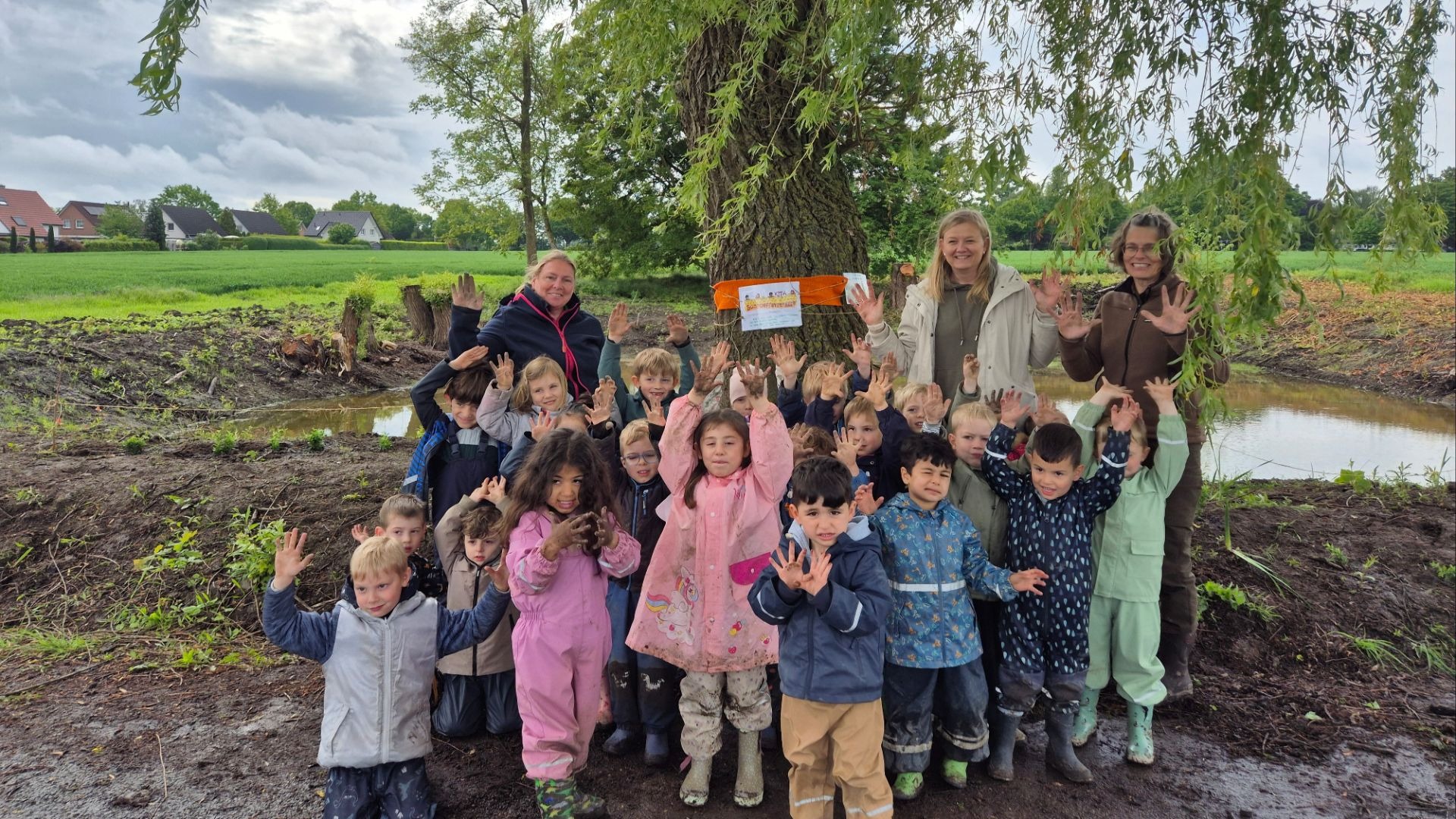 Viele lachende Kita-Kinder und drei Erwachsene zeigen ihre schmutzigen Hände vor einem großen Baum und einem Graben