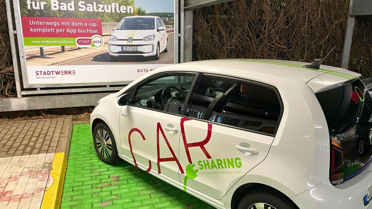 Weißes E-Auto mit "CAR SHARING" Beschriftung und grünen Streifen steht vor einem Plakat für Stadtwerke Bad Salzuflen