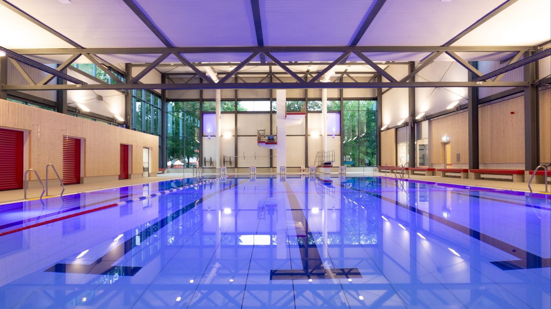 Innenansicht des Lohfeld Bades mit blau leuchtendem Schwimmbecken, Bahnen und Sprungturm