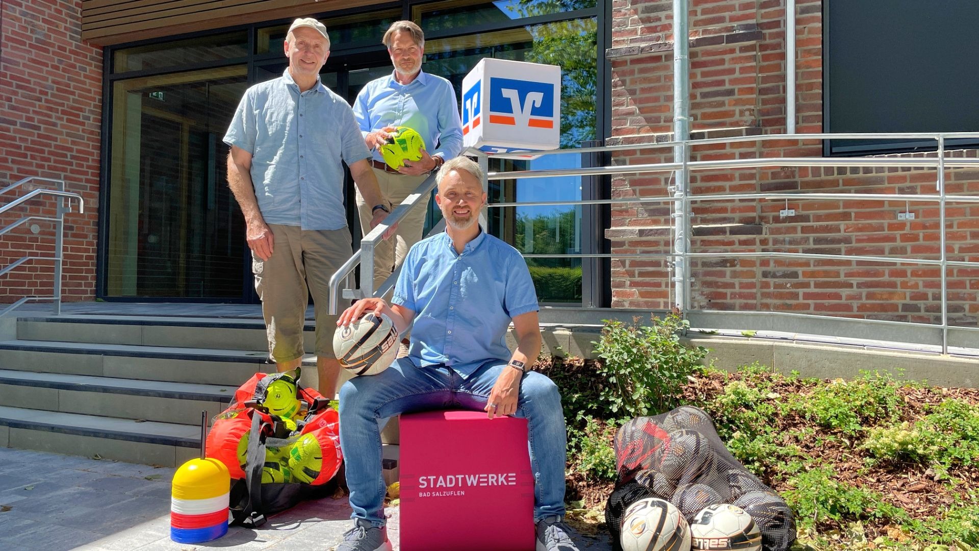 Drei Männer mit Fußbällen und Trainingsmaterialien vor einem Gebäude, auf Kisten sind Logos von Stadtwerke und Volksbank