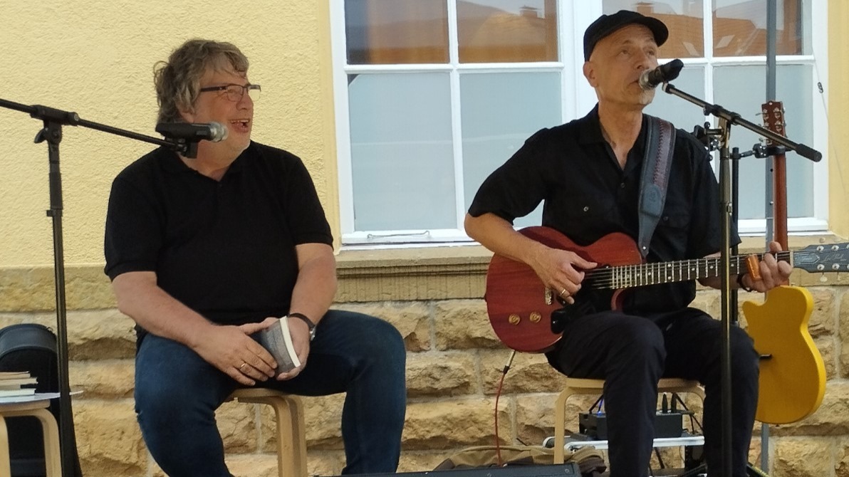Greyhound George spielt Gitarre, während Hellmuth Opitz am Mikrofon sitzt und spricht