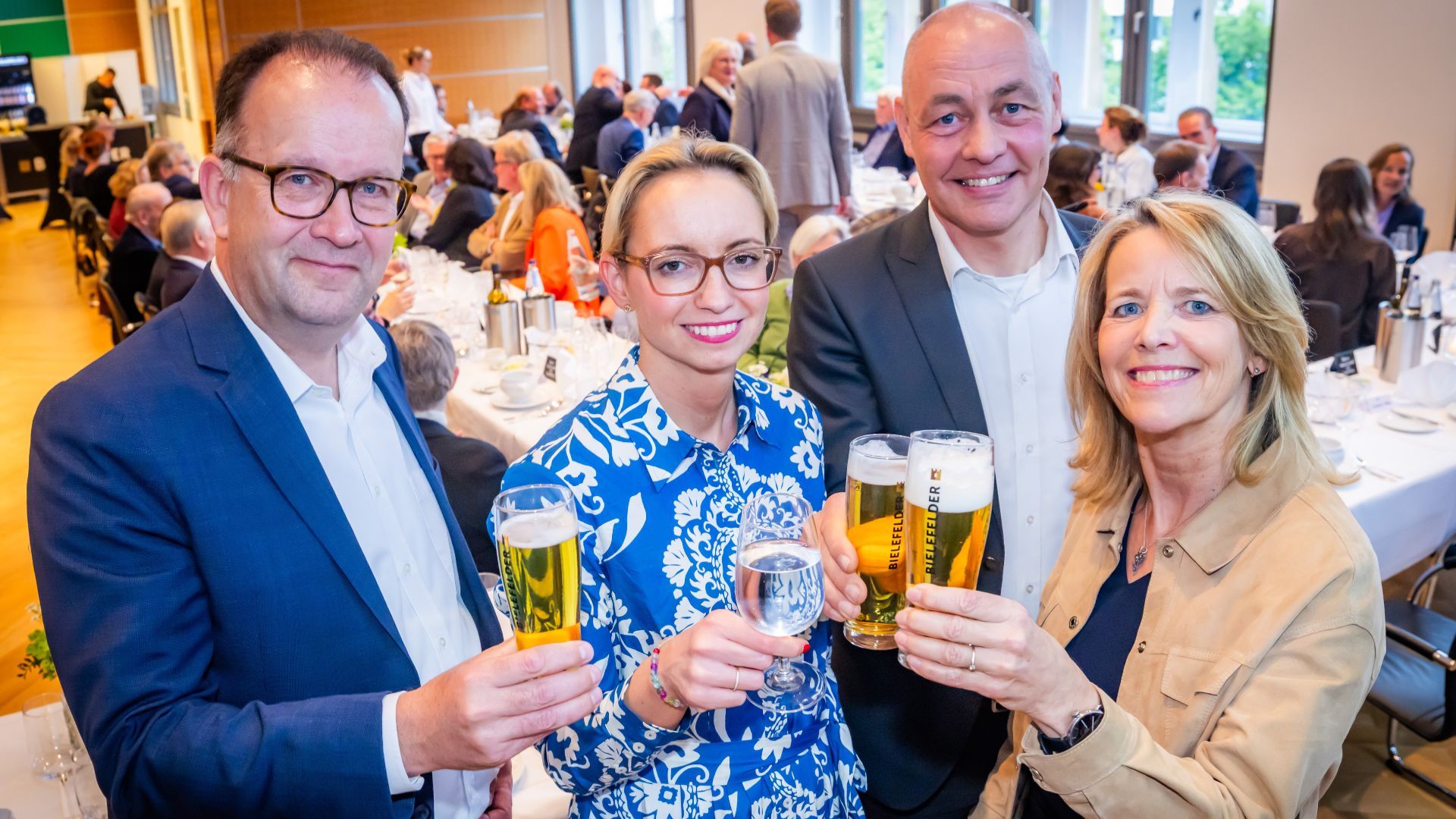 Vier lächelnde Personen stoßen bei einem Empfang mit Bielefelder Bier und Wasser an, im Hintergrund Gäste