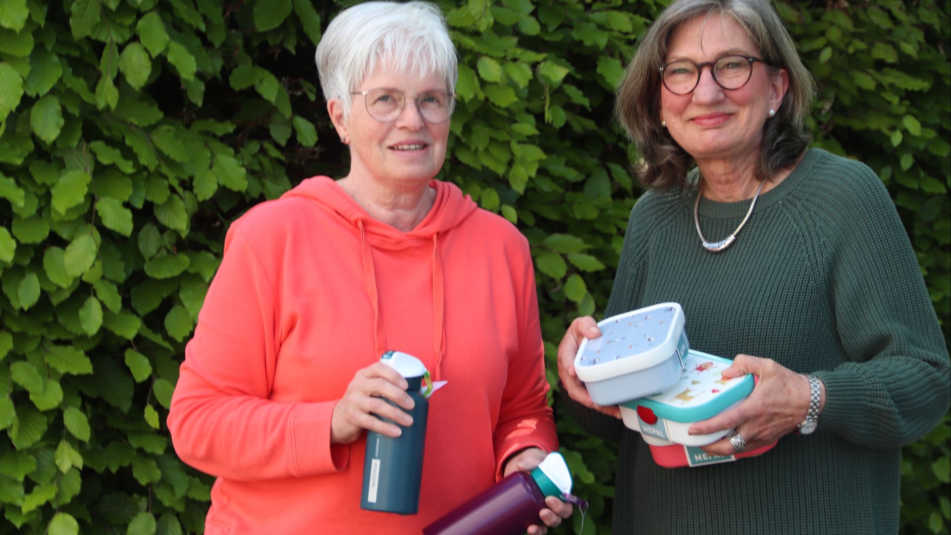 Zwei Frauen halten Trinkflaschen und bunte Brotdosen vor einer grünen Hecke