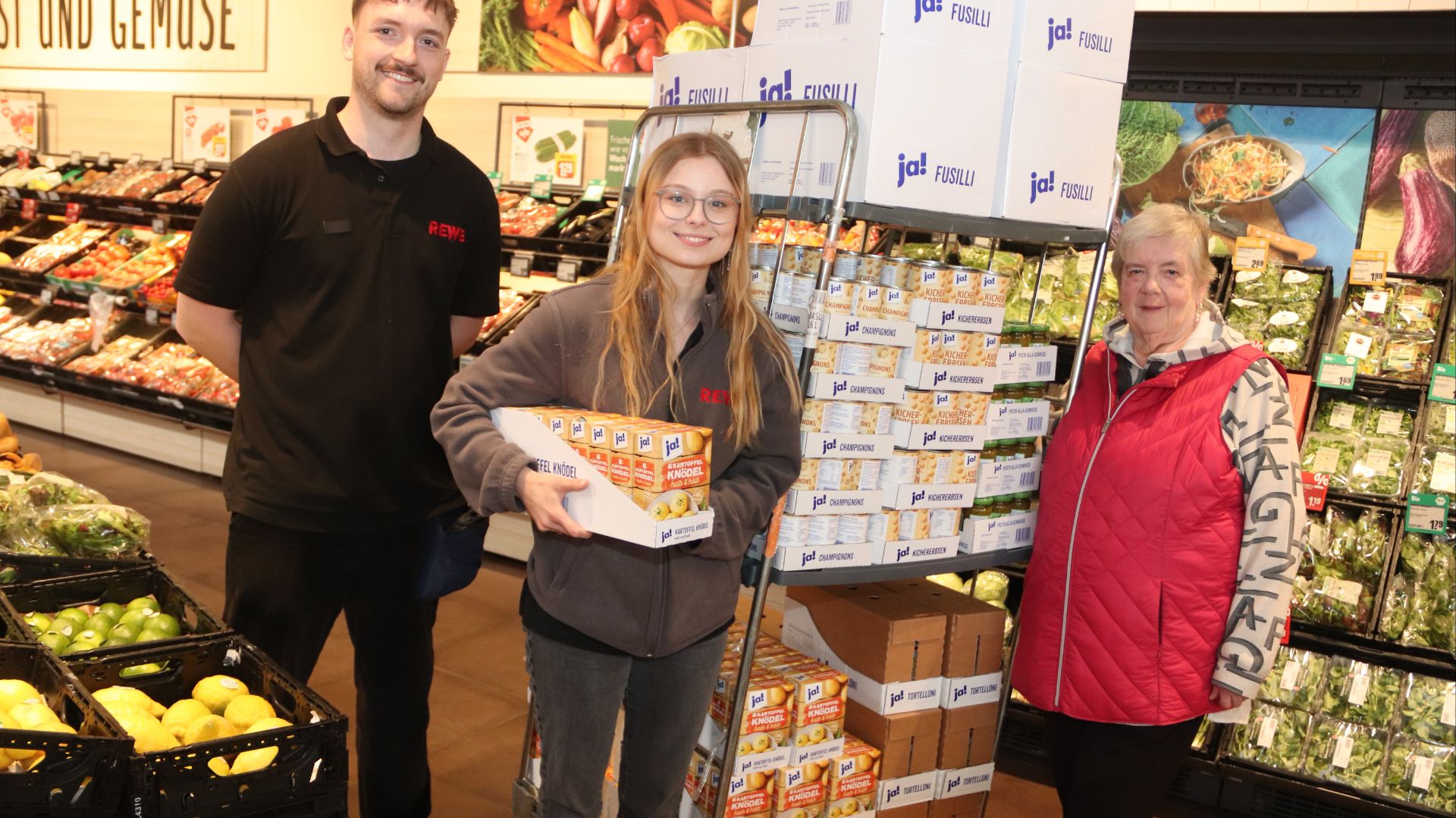 Drei Personen, darunter zwei Rewe-Mitarbeiter, stehen lächelnd neben einem Spendenwagen voller ja!-Produkte im