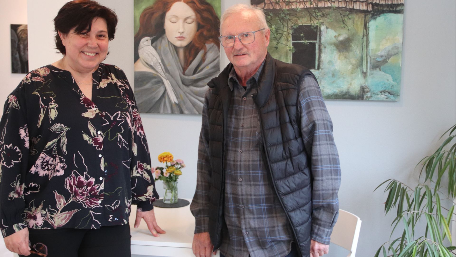 Die lächelnde Frau und der Künstler Sigurd Schade mit Brille vor seinen Gemälden und einem Blumenstrauß