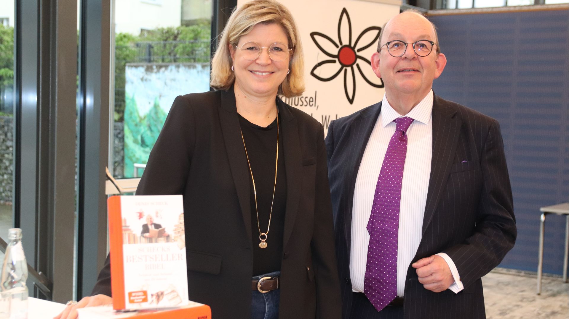 Denis Scheck und eine blonde Frau lächeln hinter einem Tisch mit seinem Buch "SCHECKS BESTSELLER BIBEL