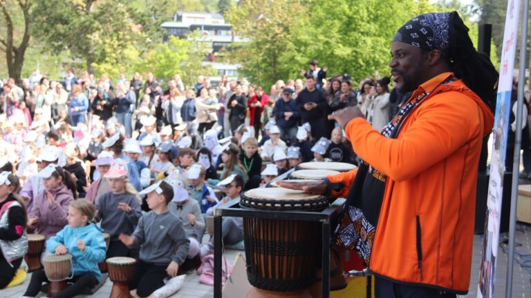 Ein Mann mit Dreadlocks und orangefarbener Jacke spielt Djembe-Trommeln vor Kindern, die auch trommeln