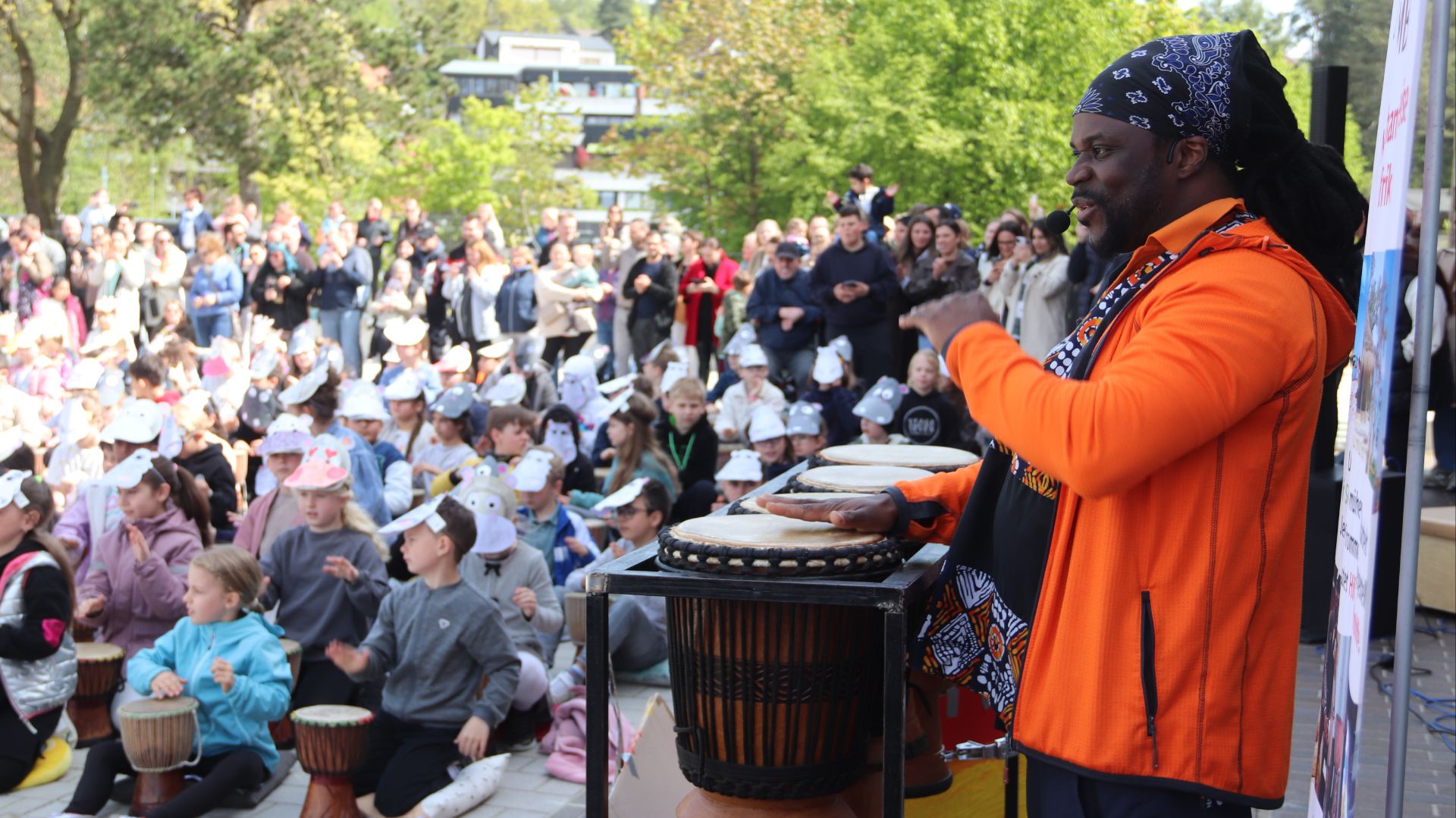 Ein Mann mit Dreadlocks und orangefarbener Jacke spielt Djembe-Trommeln vor Kindern, die auch trommeln
