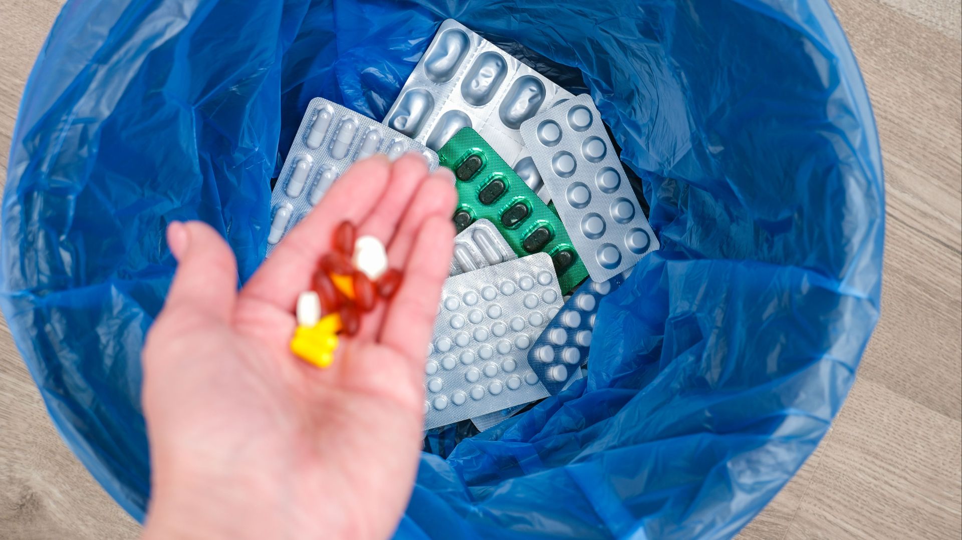 Hand hält bunte Tabletten über blauem Müllsack mit vielen Blisterverpackungen