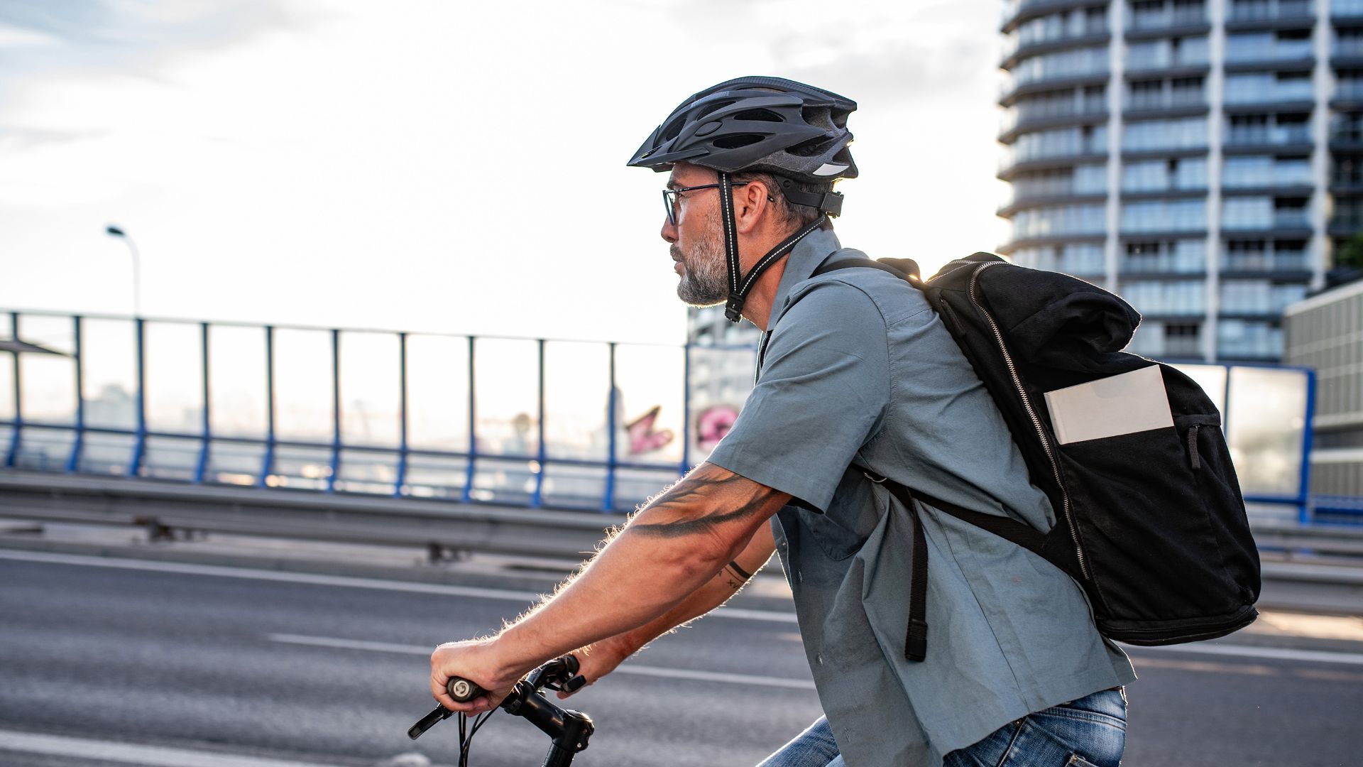 Bärtiger Mann mit Helm und Rucksack fährt auf Brücke Fahrrad, im Hintergrund ein Hochhaus