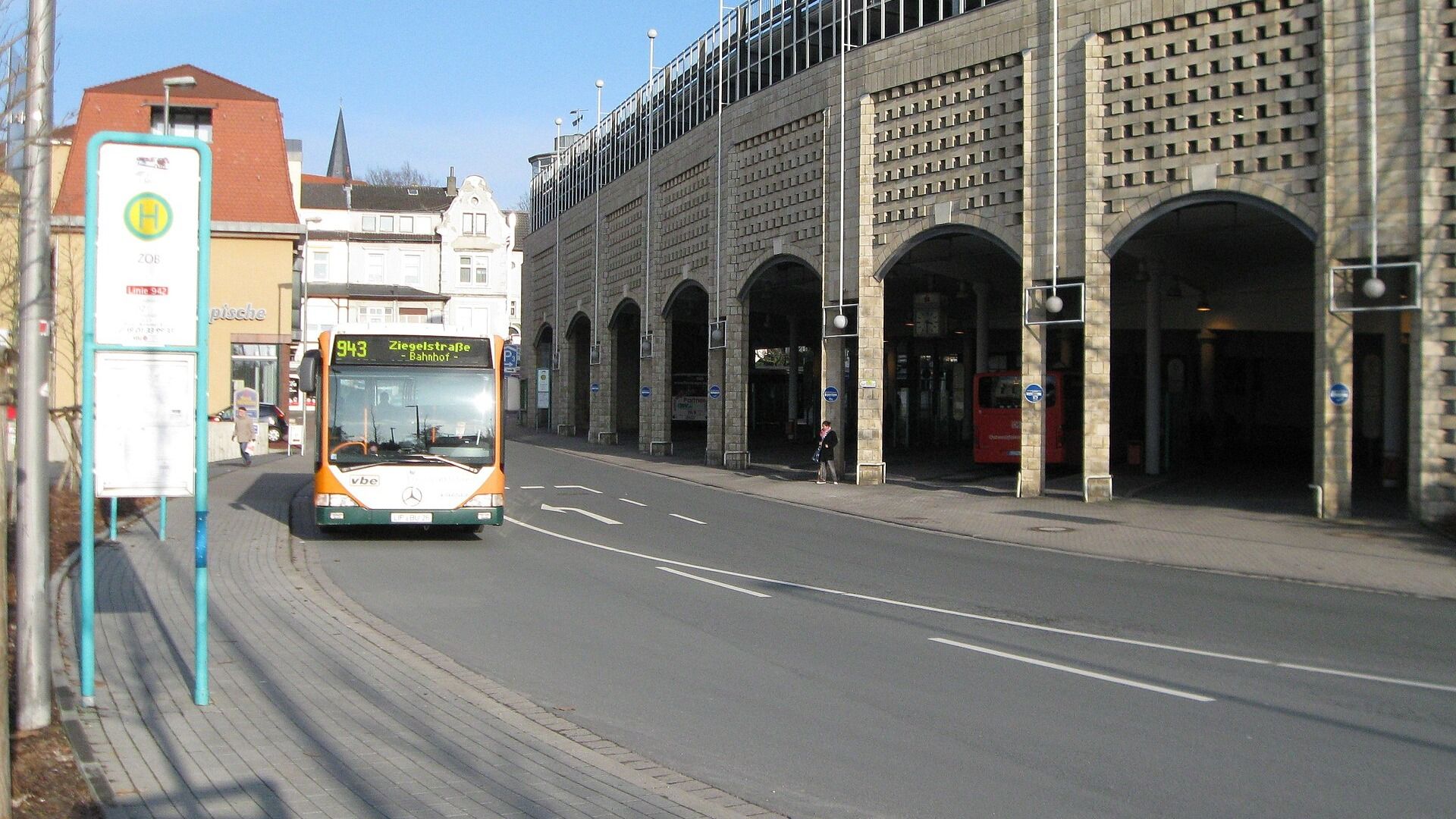 Orange-grüner Bus Linie 943 fährt auf einer Straße an einer Haltestelle und einem großen Gebäude mit Bögen vorbei