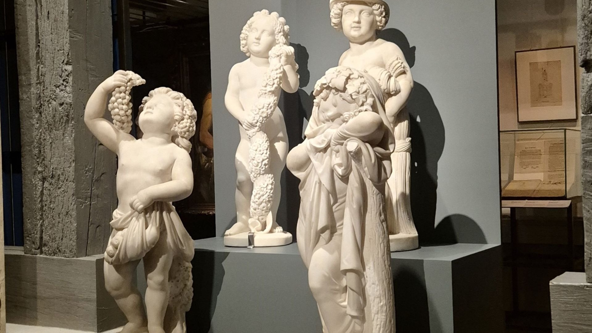 Vier weiße Marmorskulpturen von Kindern mit Trauben, Girlanden und Tüchern, auf grauen Podesten im Museum