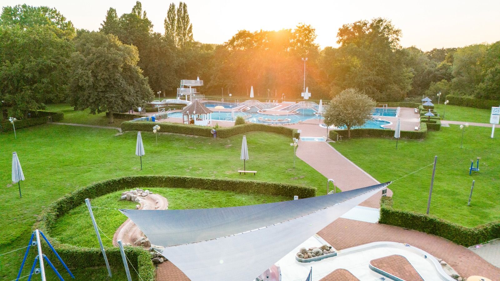 Luftaufnahme: Freibad mit mehreren Becken, Wasserrutsche, großen Liegewiesen und Bäumen im warmen Sonnenlicht