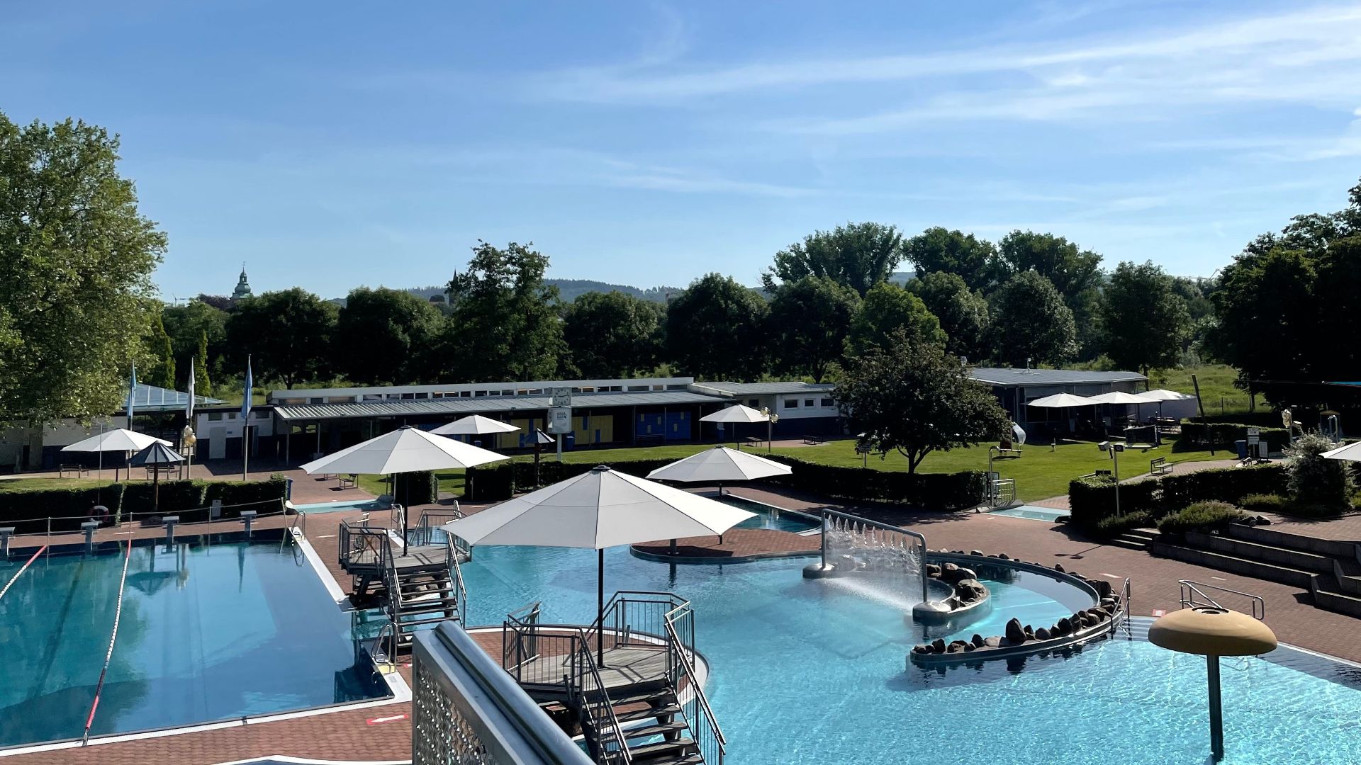 Ein leeres Freibad mit mehreren Schwimmbecken, Sonnenschirmen, Gebäuden und grünen Bäumen unter blauem Himmel
