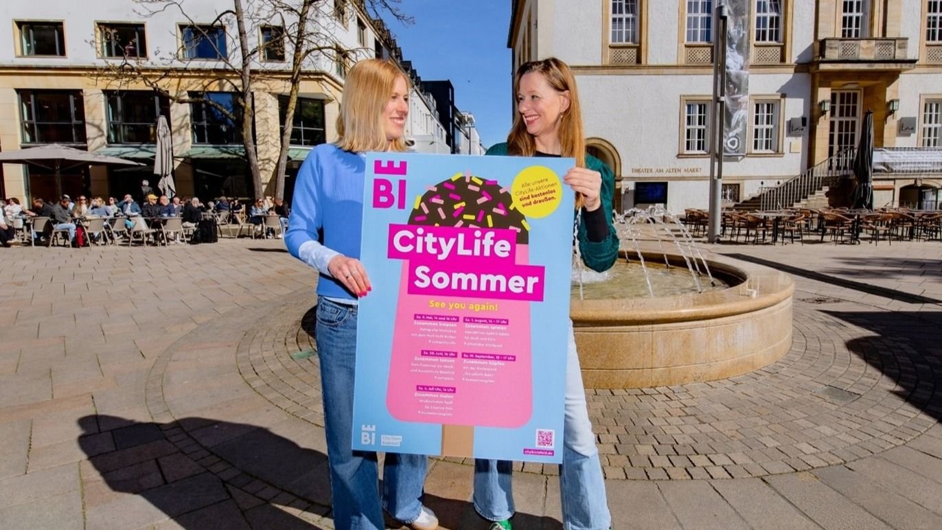 Zwei Frauen lächeln sich an, halten ein blaues Plakat „CityLife Sommer BI“ vor einer Caféterrasse und einem Brunnen