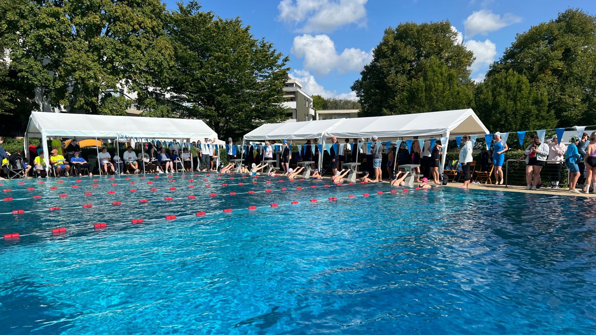 Schwimmer stehen an Startblöcken im Freibad, Zuschauer und Betreuer unter weißen Zelten am Beckenrand bei einem
