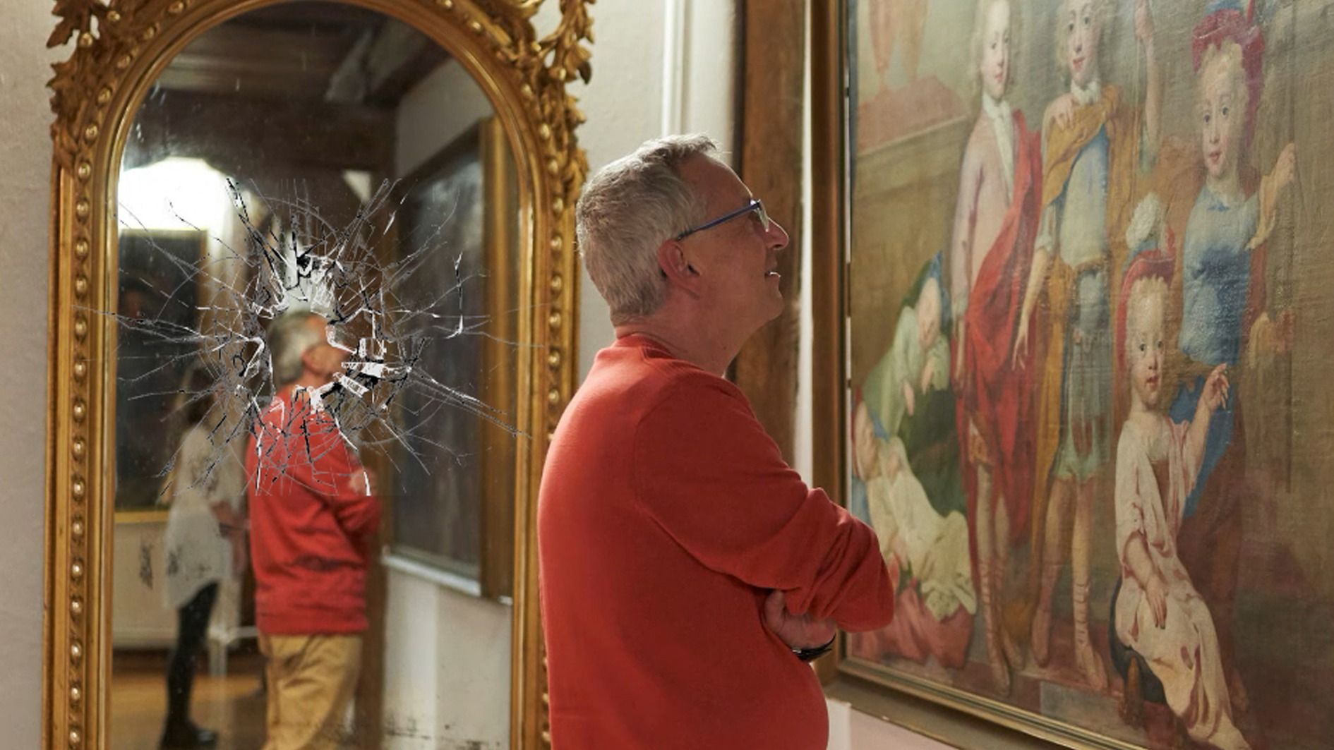 Mann in orangefarbenem Hemd blickt auf ein Gemälde, daneben ein goldener Spiegel mit zerbrochenem Glas