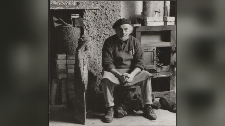 Künstler Bruno Buschmann sitzt in seinem Atelier, trägt eine Schürze und Baskenmütze, neben einer Skulptur und Ofen
