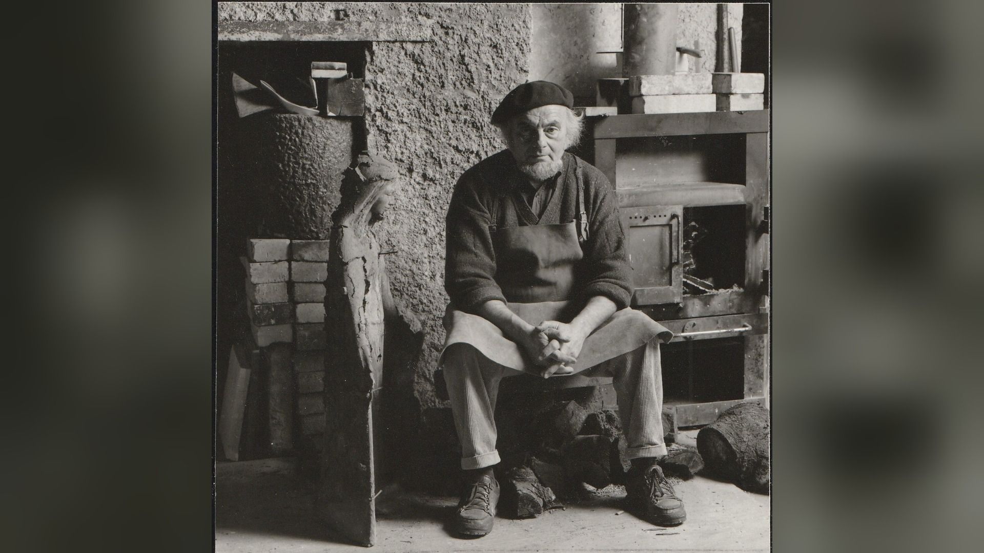 Künstler Bruno Buschmann sitzt in seinem Atelier, trägt eine Schürze und Baskenmütze, neben einer Skulptur und Ofen