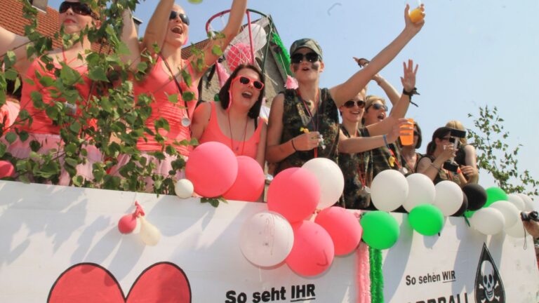 Lachende Frauen in bunten Oberteilen und Tarnkleidung feiern auf einem mit Ballons und grünen Zweigen geschmückten Wagen