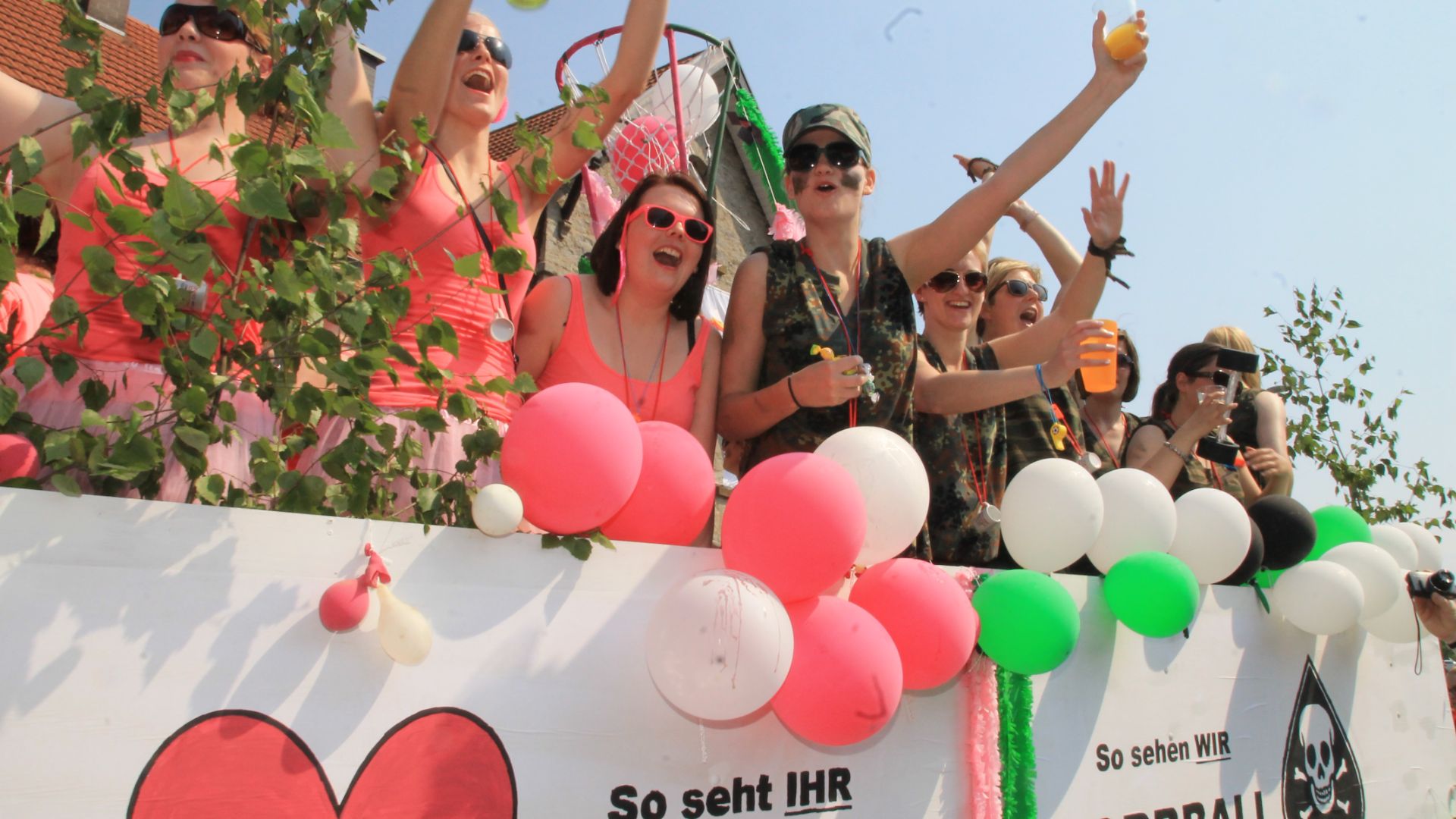 Lachende Frauen in bunten Oberteilen und Tarnkleidung feiern auf einem mit Ballons und grünen Zweigen geschmückten Wagen