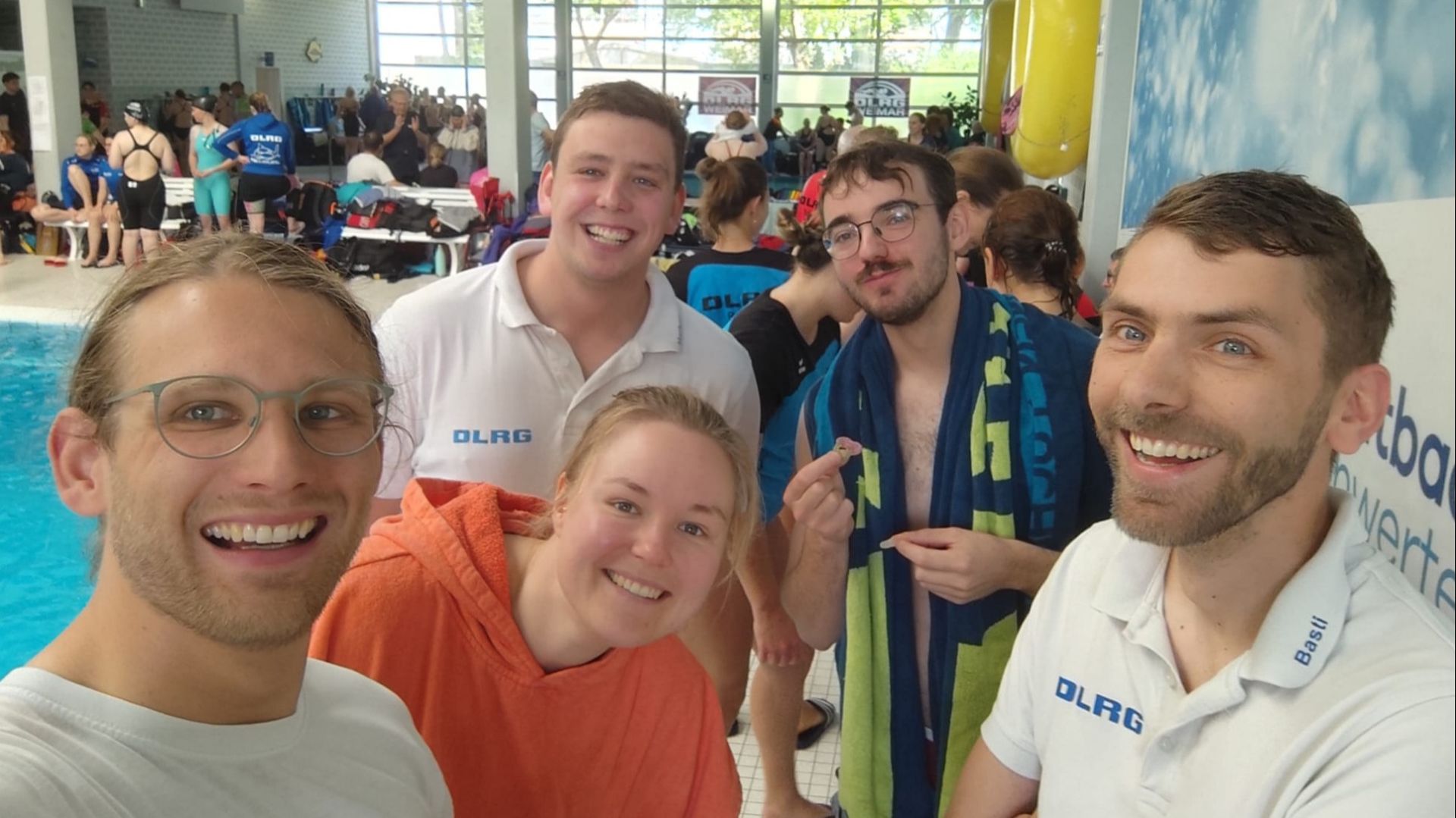 Fünf DLRG-Mitglieder lächeln in einem Schwimmbad in die Kamera, im Hintergrund weitere Menschen am Beckenrand