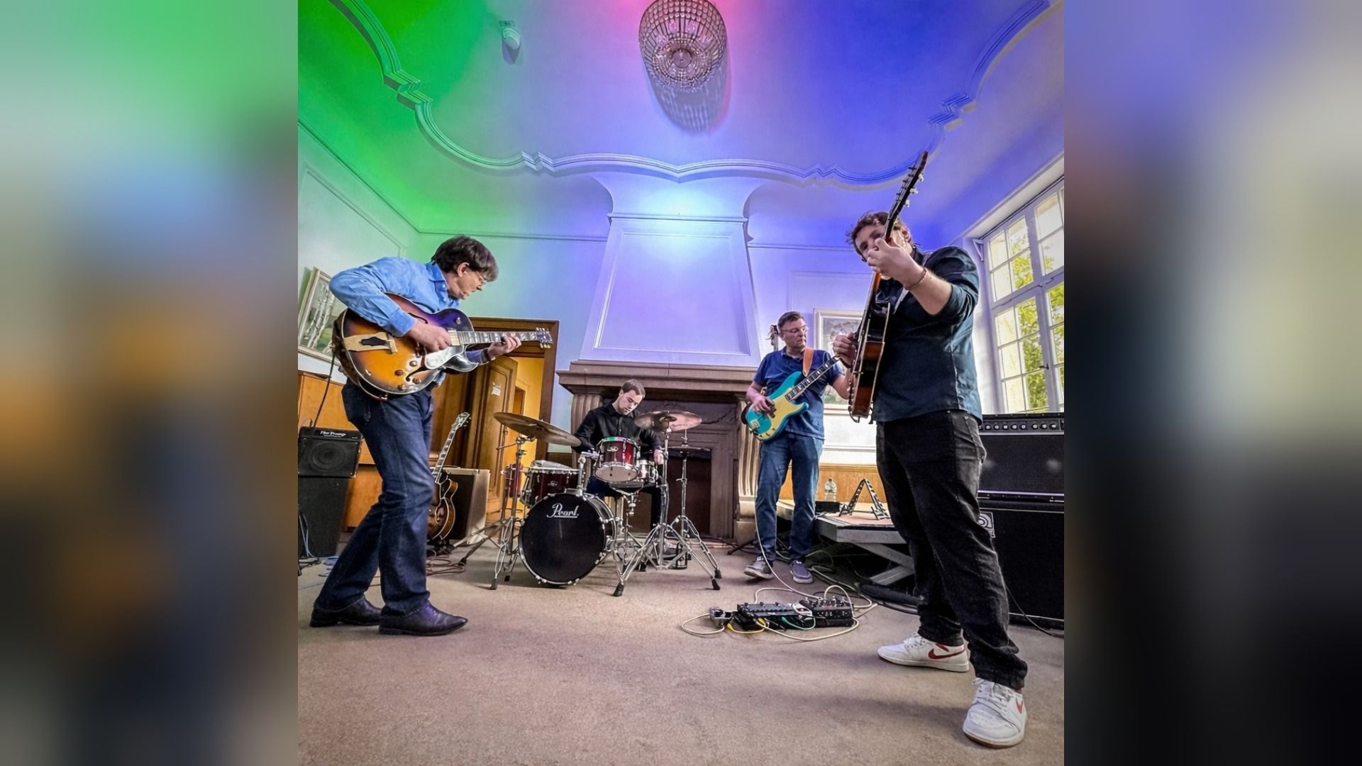 Vier Musiker spielen Gitarre, Bass und Schlagzeug in einem prächtigen Raum mit farbig beleuchteter Decke und Kamin