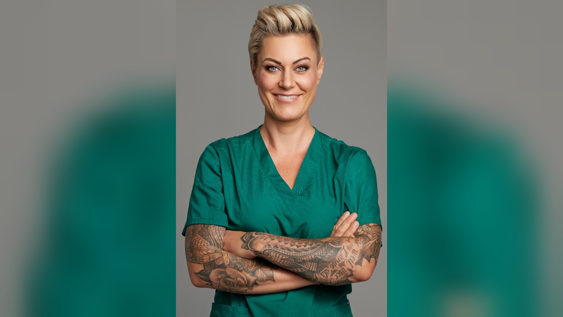Doc Caro lächelt in grüner Arztkleidung mit kurzen blonden Haaren und verschränkten, tätowierten Armen