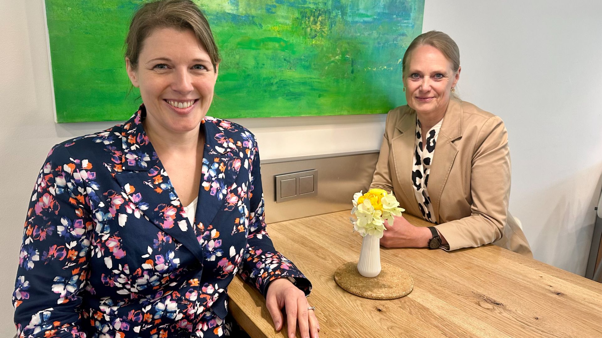 Zwei Frauen sitzen lächelnd an einem Holztisch, dazwischen eine Vase mit gelben Blumen