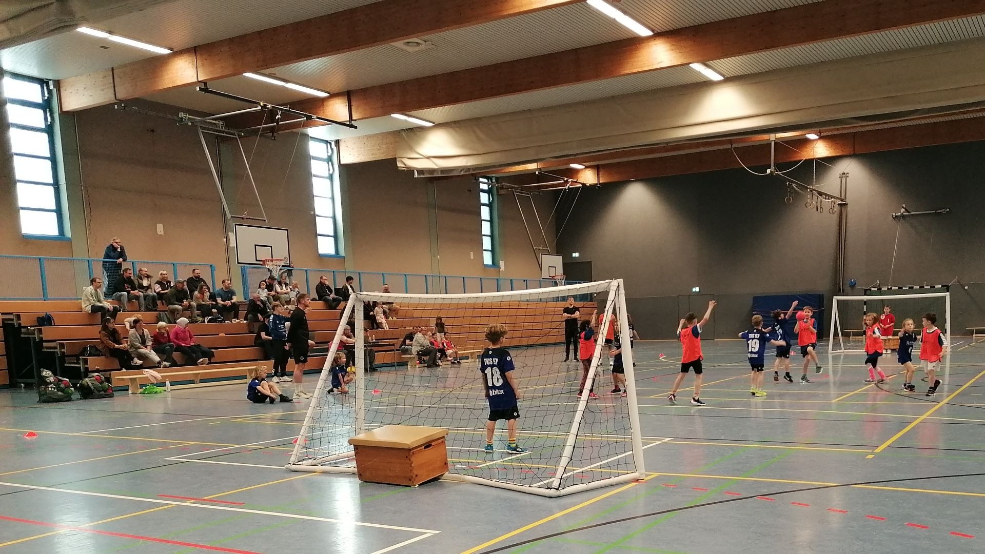 Mehrere Kinder in Trikots spielen Handball in einer Sporthalle mit Zuschauern auf der Tribüne und zwei Toren