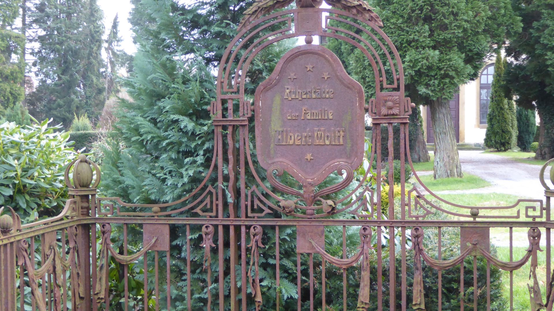 Rostiges verziertes Eisentor mit Inschrift "Ruhestätte der Familie Albert Wolff" auf einem historischen Friedhof mit