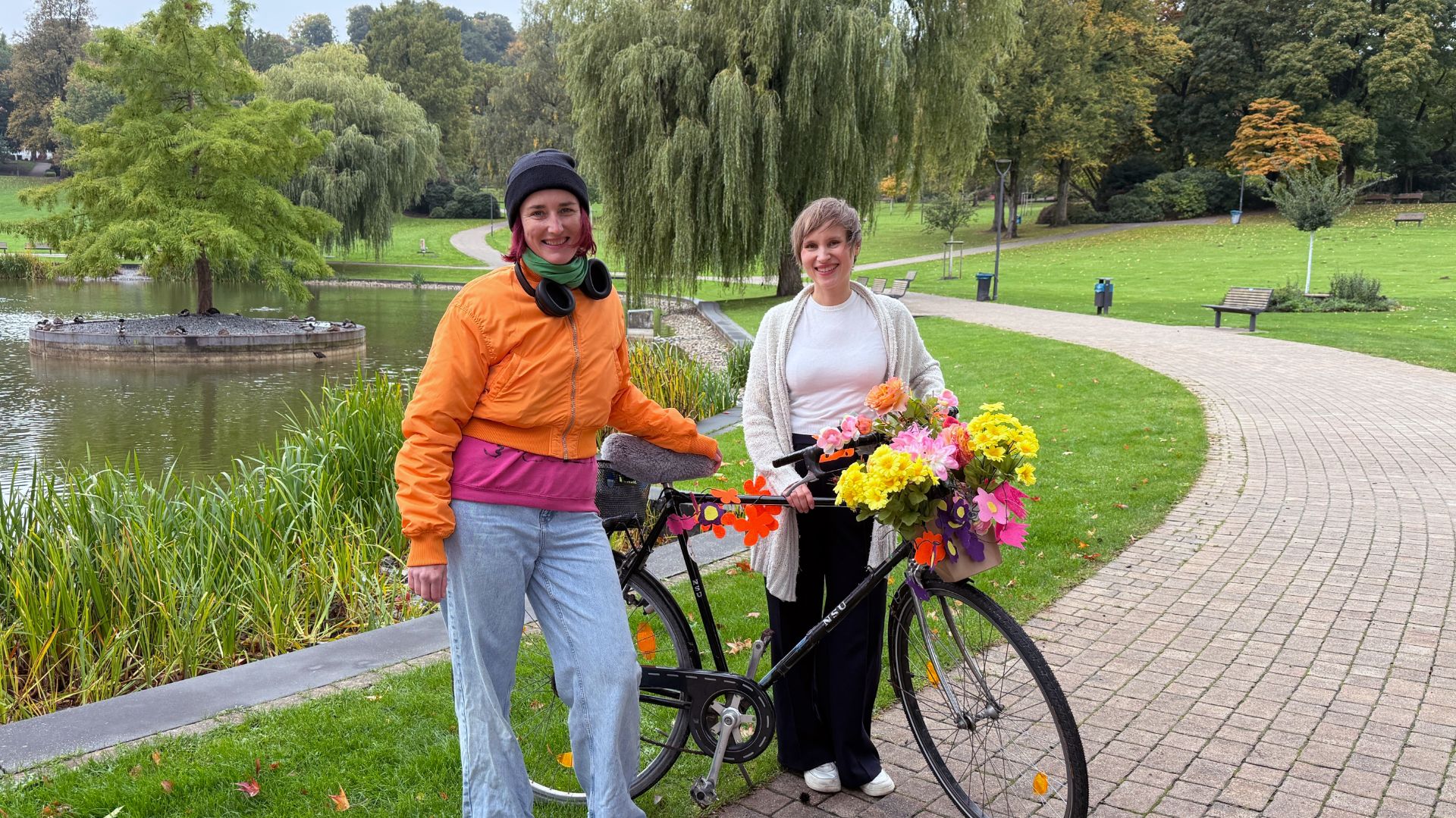 Zwei lächelnde Frauen stehen neben einem mit bunten Blumen verzierten Fahrrad in einem Park mit Teich und Bäumen